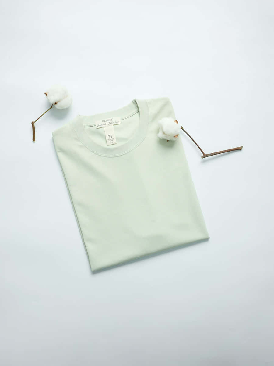 Camiseta de algodón verde salvia doblada sobre fondo blanco, con ramas de algodón al lado