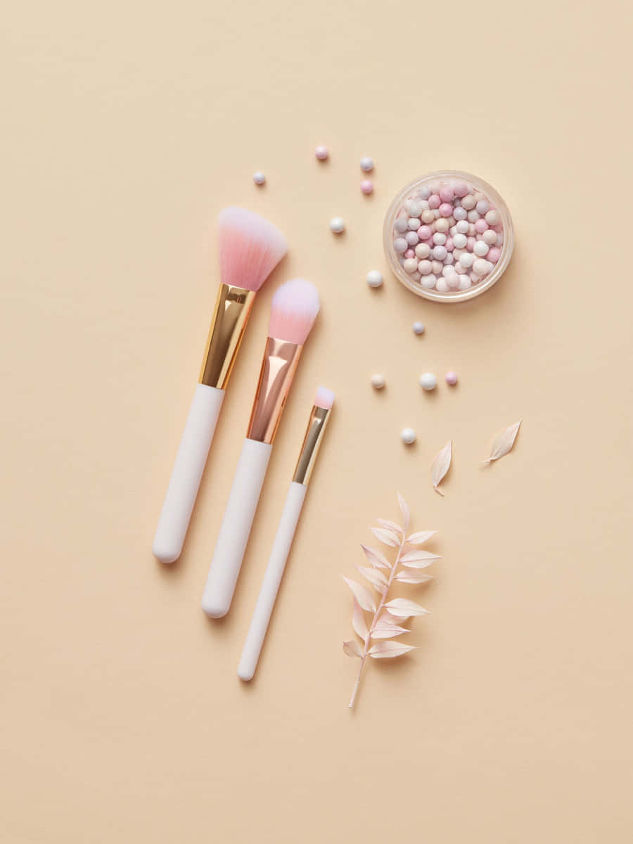 Tres brochas de maquillaje de mango blanco y virola dorada sobre fondo beige, junto a un frasco de perlas de rubor